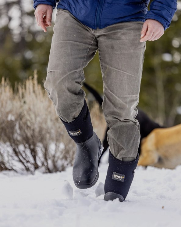 The image shown is of the Men's Bozeman tall insulated winter boots.  The man is walking through the snow with his dogs in the background.