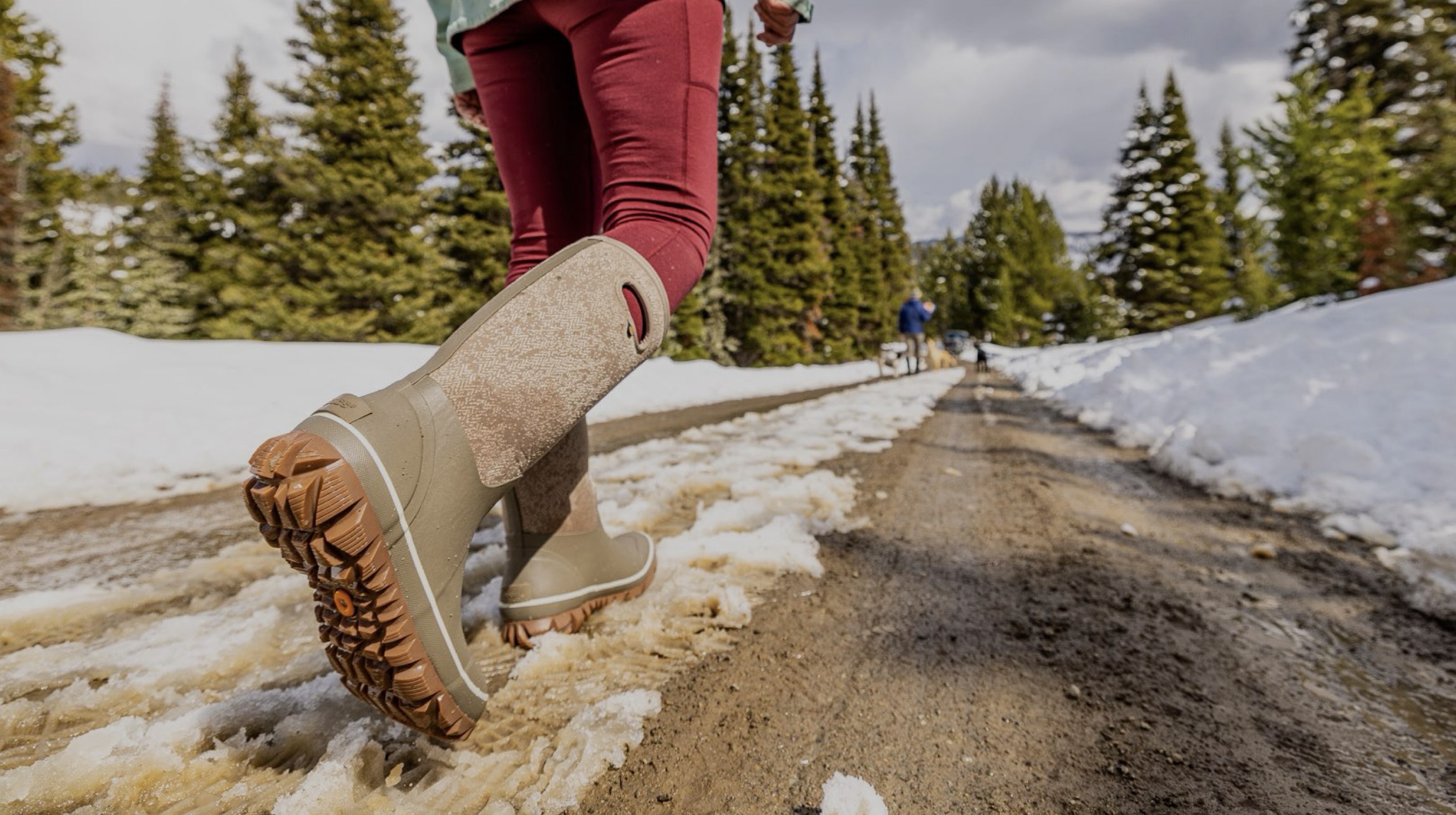 The image shown is of the Whiteout Faded tall insulated winter boots in a tan print.  The woman is walking through the snow with his dogs in the background.