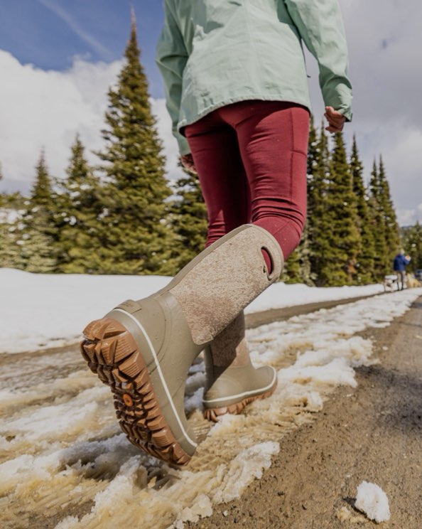 The image shown is of the Whiteout Faded tall insulated winter boots in a tan print.  The woman is walking through the snow with his dogs in the background.