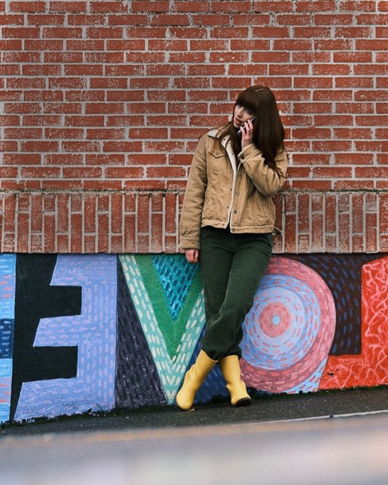 Shop our Women's New Arrivals.  The image shown is a woman leaning back on a brick wall, talking on the phone.  The boot highlighted our Women's Amanda Mid rainboots in yellow.