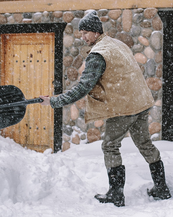 The image shown is a man shoveling snow outside of their cabin.  Shop our top Men’s winter boots. 