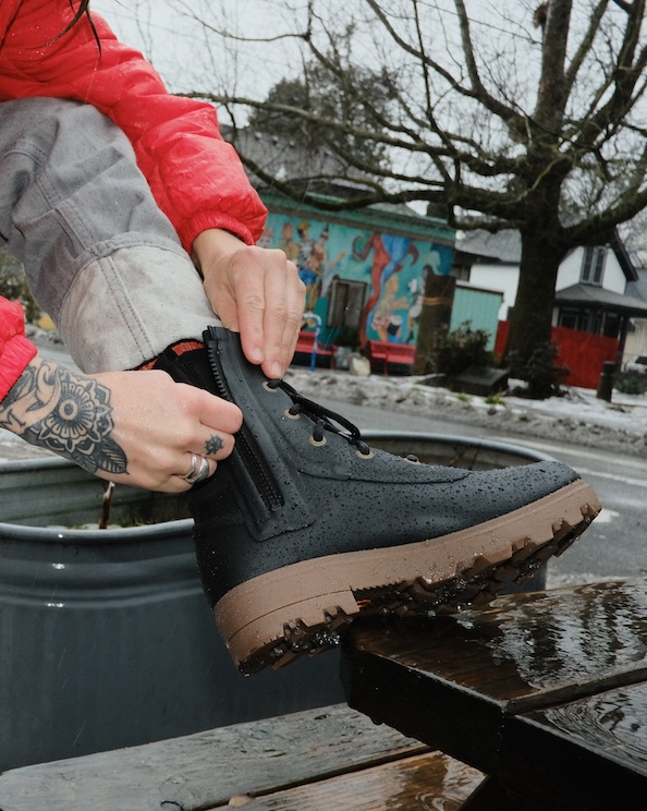 The image shown is a woman in a red jacket zipping up her Holly Rain Lace boots in black.  The backdrop is on a winter urban street. 