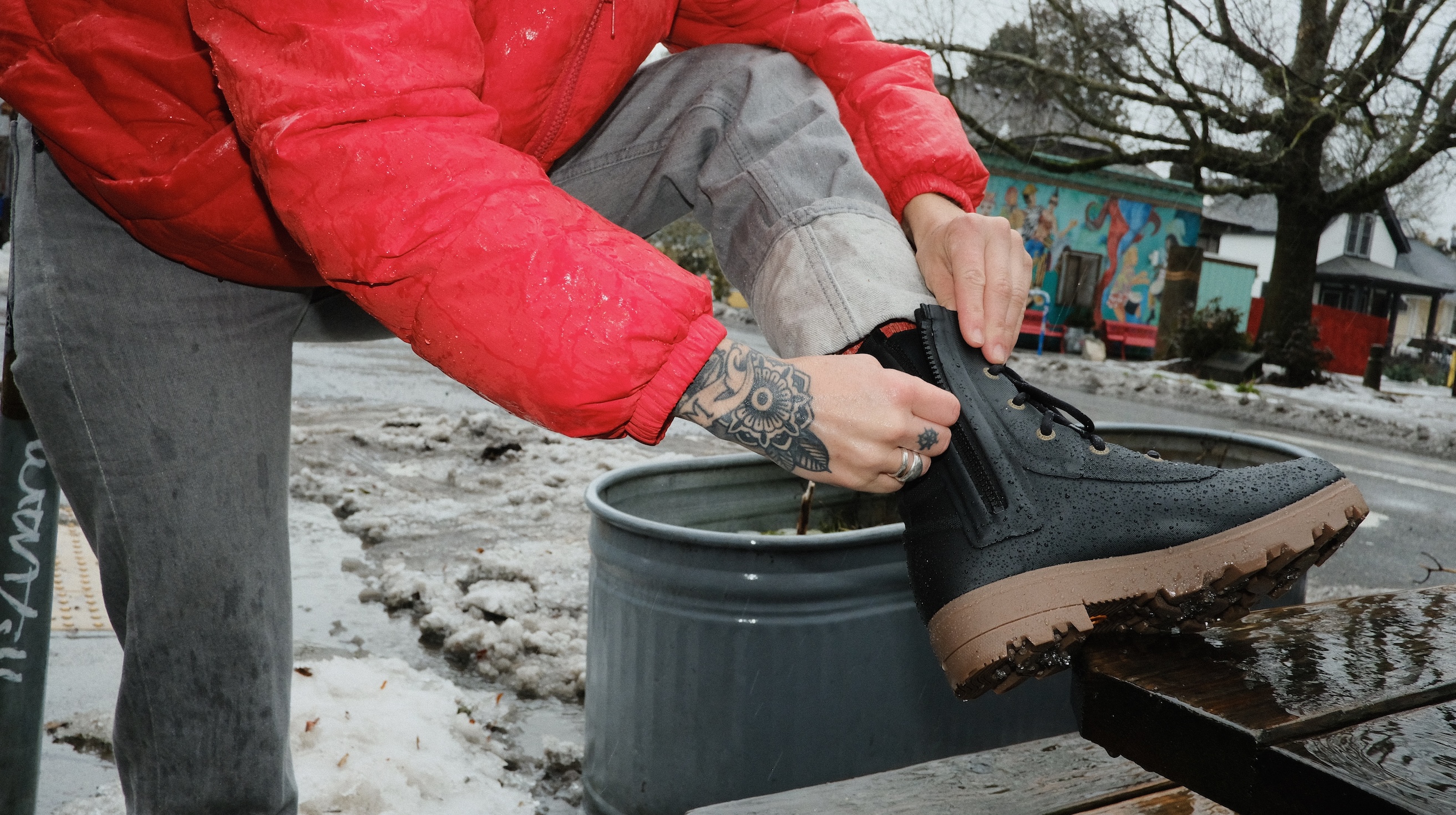 The image shown is a woman in a red jacket zipping up her Holly Rain Lace boots in black.  The backdrop is on a winter urban street. 