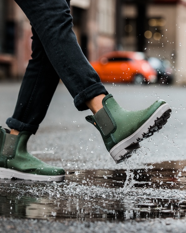 The image shown is a closeup of our women’s holly rainboot Chelsea in green splashing through a puddle.  Shop now.