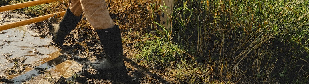 The image shown is our Men's Classic Seamless tall waterproof work boot in black pushing open a gate. Shop now. 